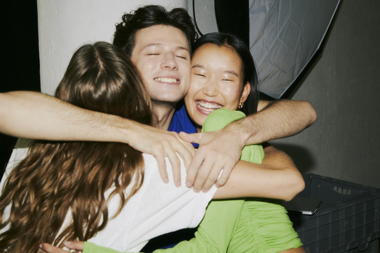 Three young people laugh and smile as they share a group hug, showing joy and friendship. One person is facing away, while the other two smile brightly at the camera.