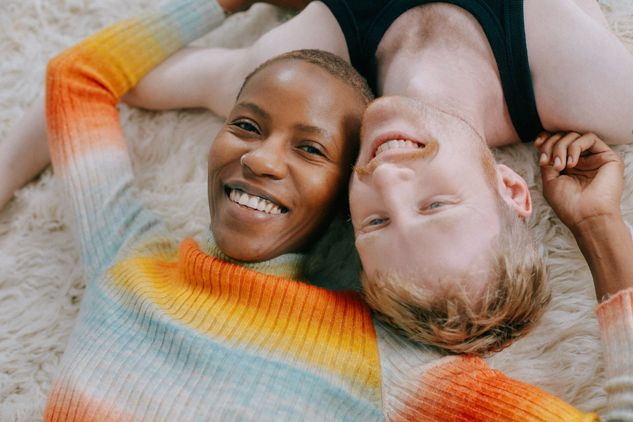 Two people lie on a soft, light rug, smiling and holding hands. One wears a colorful striped sweater, the other a black top. Their heads are close together, creating a warm, joyful atmosphere.