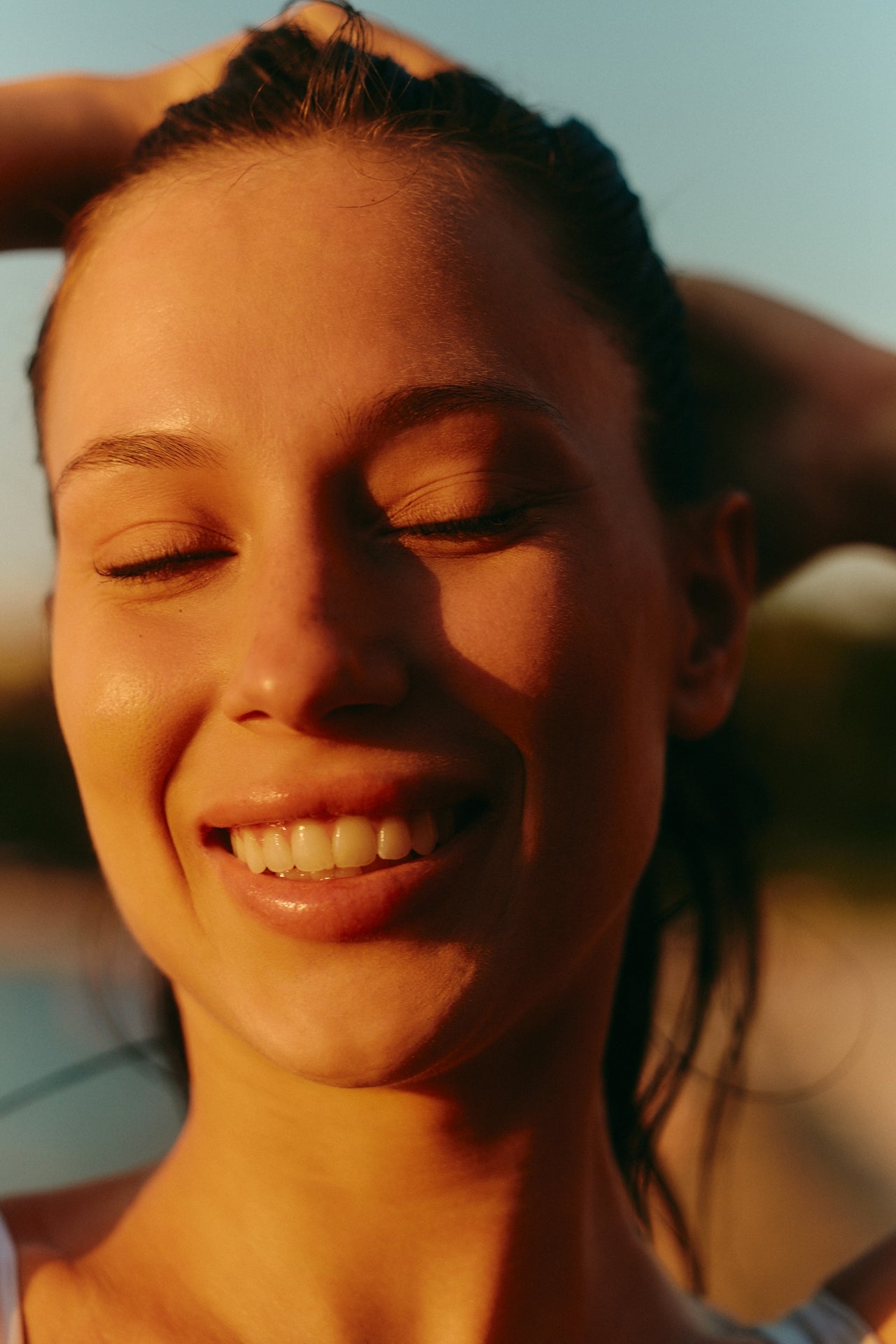 A woman with wet hair smiles with her eyes closed, bathed in warm sunlight. She has her hands behind her head, and the background is softly blurred, suggesting an outdoor setting.